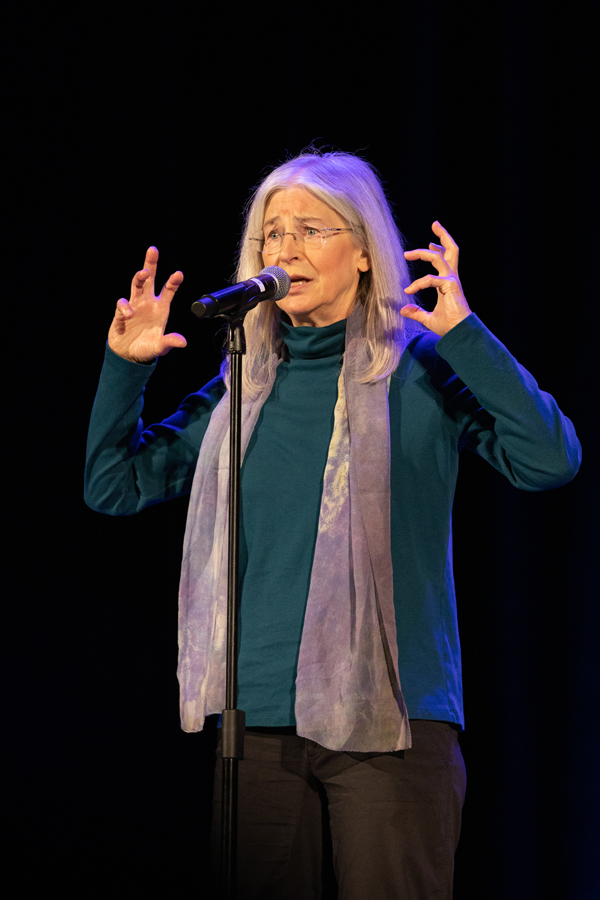 The image depicts a woman with shoulder length gray hair wearing a turquoise sweater and a gray scarf and glasses sharing a story in front of a microphone. The photo was taken by kmr studios.
