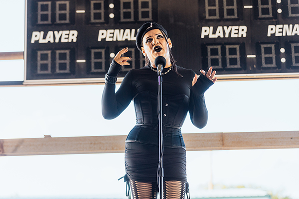 The image shows a woman holding her hands up as she shares a story into a microphone. She is standing in front of a hockey scoreboard. Photo credit goes to Donal Lakatua.