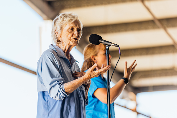 The image shows a woman standing at a microphone sharing a story. To her left, another woman stands providing American Sign Language interpretation. The woman is wearing blue clothing. Photo credit: Donal Lakatua.