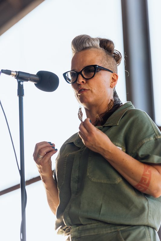 The photo features a woman wearing glasses and sharing a story into a microphone at an outdoor event. Photo credit: Donal Lakatua.