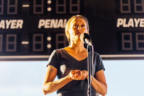 The image shows a woman sharing a story into a microphone during a performance. She is standing in front of a scoreboard at an arena. The photo was taken by Donal Lakatua.