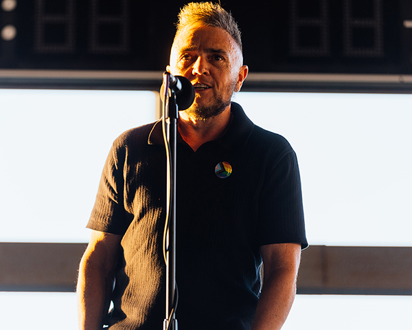 The image shows a man sharing a story into a microphone during a performance. He is standing in front of a scoreboard at an arena. The photo was taken by Donal Lakatua.