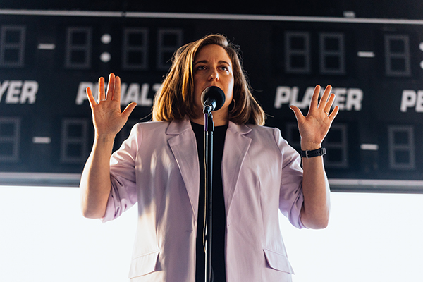 The image shows a woman sharing a story into a microphone during a performance.  She is standing in front of a scoreboard at an arena. The photo was taken by Donal Lakatua.