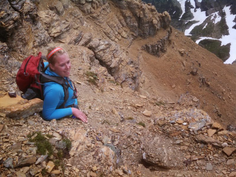 The image shows a woman wearing a backpack while standing on a rocky hillside looking up at the camera. The setting is outdoors in a mountainous or hilly area.