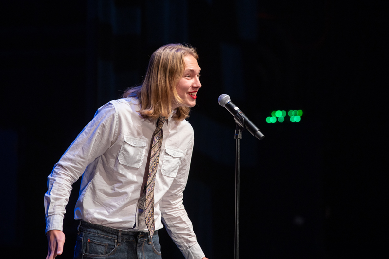 The image depicts a person speaking into a microphone on stage. The person is wearing jeans and is standing near a microphone stand.