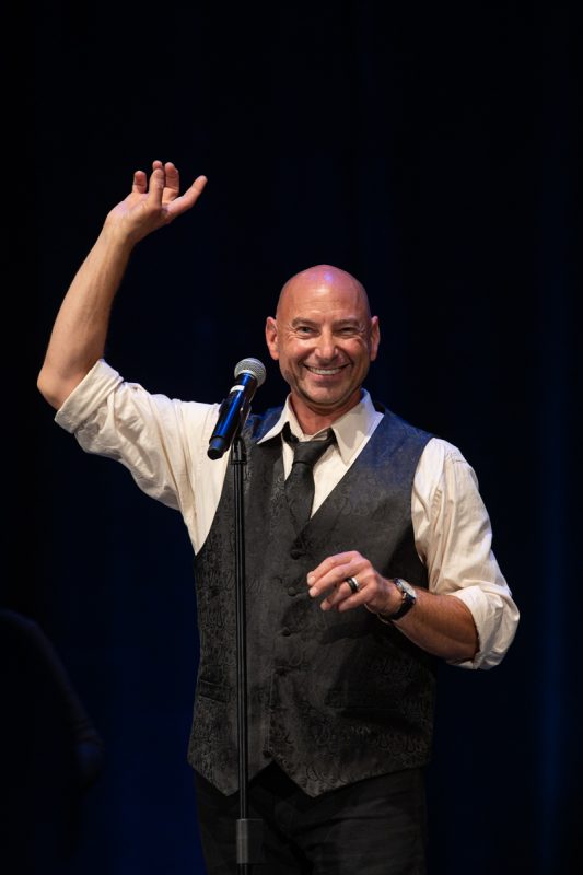 The image depicts a smiling man holding a microphone and raising his hands. He is wearing a dress shirt and vest.